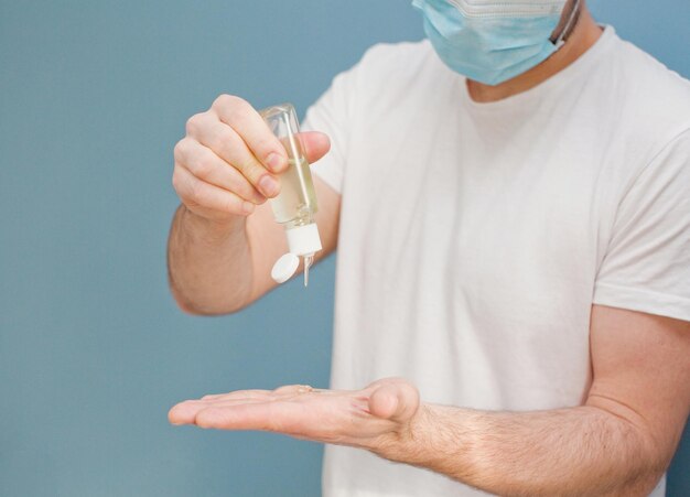 Joven con mascarilla quirúrgica usa desinfectante de manos para limpiarse las manos de gérmenes en un fondo azul