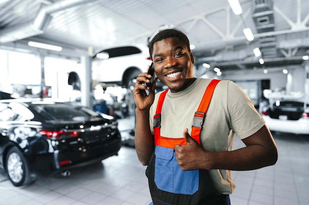 Joven mecánico africano vestido con uniforme usando teléfono móvil en un servicio de automóviles