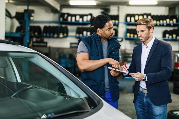 Un joven mecánico de automóviles negro y su gerente se comunican mientras revisan la lista de verificación en un taller de reparación