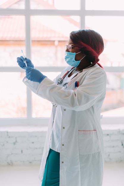 Joven médico afroamericano sonriente en máscara médica sosteniendo una jeringa