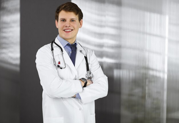 Foto un joven médico alegre está de pie con los brazos cruzados en una clínica. retrato de un médico profesional en el trabajo. concepto de medicina.