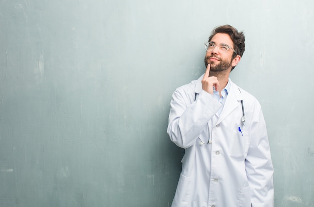 Un joven médico amigable contra una pared de grunge con un espacio de copia pensando y mirando hacia arriba, confundido acerca de una idea, estaría tratando de encontrar una solución