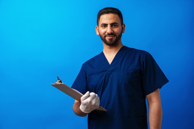 Joven médico árabe sosteniendo un portapapeles sobre un fondo azul.