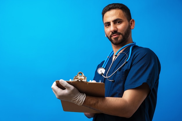 Joven médico árabe sosteniendo un portapapeles sobre un fondo azul.