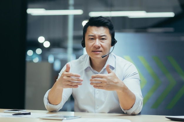 Un joven médico asiático se sienta en la oficina en una mesa con auriculares y realiza una consulta en línea
