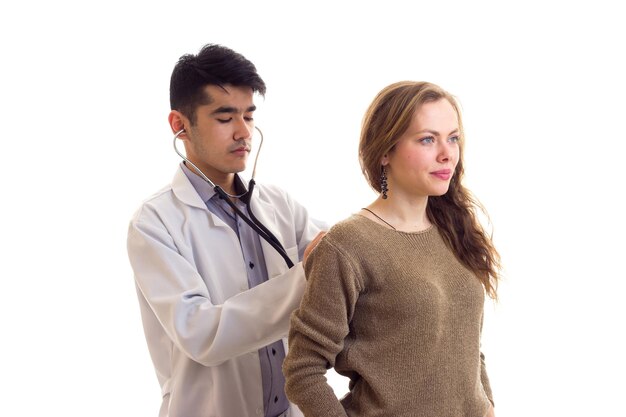 Joven médico atractivo con vestido blanco con estetoscopio escuchando la espalda de una joven agradable