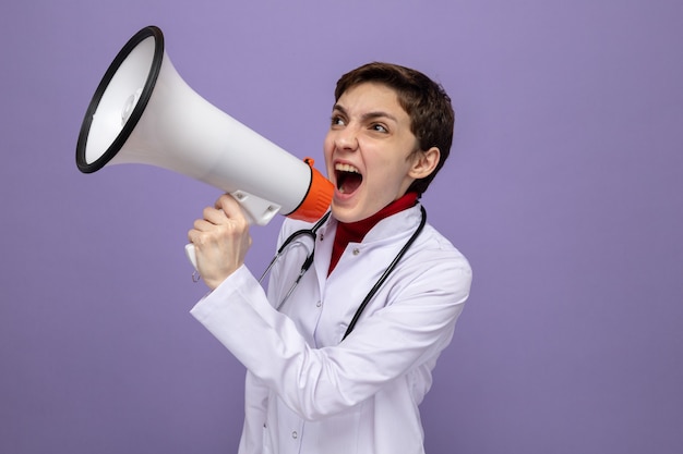 Joven médico en bata blanca con estetoscopio alrededor del cuello gritando al megáfono enojado y emocionado