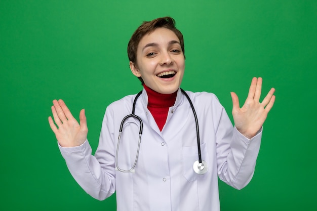 Joven médico en bata blanca con estetoscopio alrededor del cuello mirando al frente feliz y alegre levantando los brazos sobre la pared verde