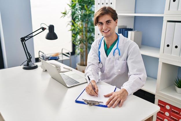 Joven médico caucásico sonriendo confiado escribiendo un informe médico en la clínica