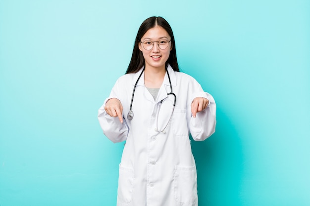 Joven médico chino mujer apunta hacia abajo con los dedos, sentimiento positivo.