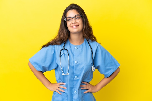 Joven médico cirujano mujer aislada sobre fondo amarillo posando con los brazos en la cadera y sonriendo