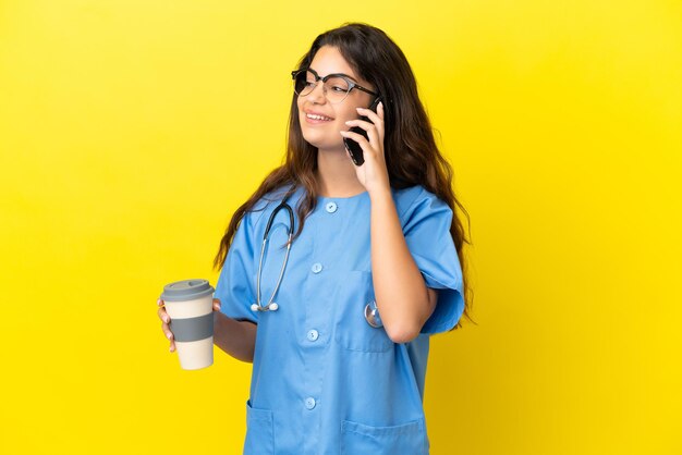 Joven médico cirujano mujer aislada sobre fondo amarillo sosteniendo café para llevar y un móvil