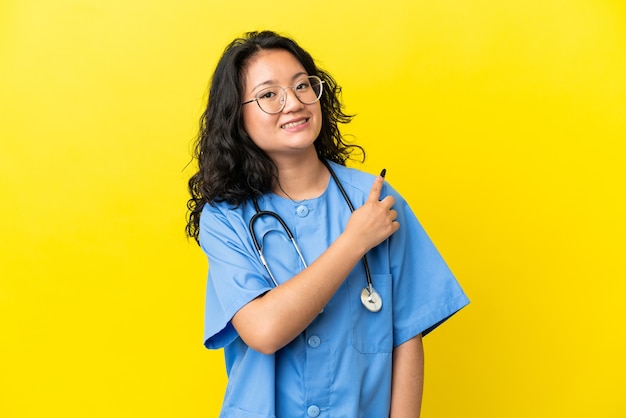 Joven médico cirujano mujer asiática aislada sobre fondo amarillo apuntando hacia el lado para presentar un producto