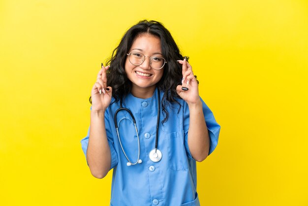 Joven médico cirujano mujer asiática aislada sobre fondo amarillo con los dedos cruzados