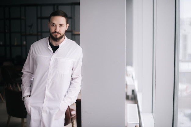 Joven médico confiado Médico caucásico con barba parado junto a la ventana y mirando a la cámara Médico masculino posando en la moderna sala de reuniones del hospital Concepto de trabajo médico