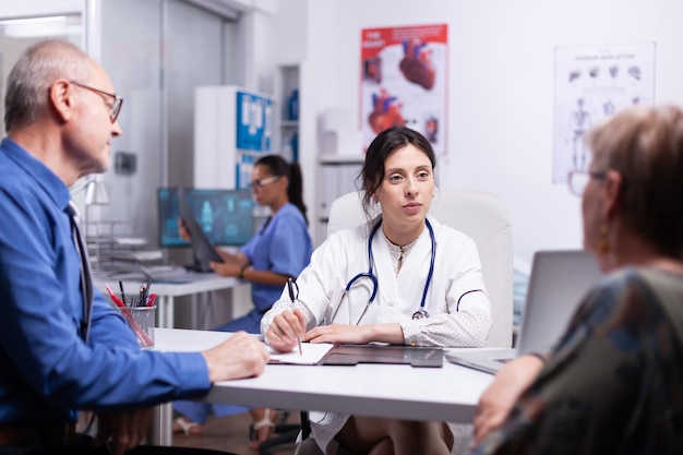 Joven médico consulta pareja de ancianos sentados en el consultorio médico, mientras que la enfermera médica trabaja en segundo plano. Médico haciendo preguntas a pacientes maduros que llenan el formulario de historial de enfermedades, prescriben