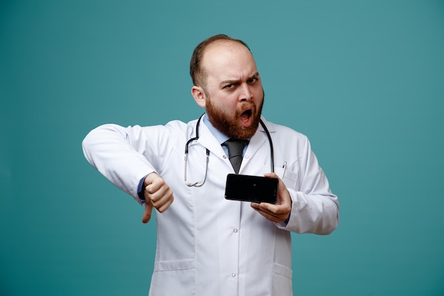 joven médico descontento con abrigo médico y estetoscopio alrededor del cuello que muestra el teléfono móvil y el pulgar hacia abajo mirando la cámara aislada en el fondo azul