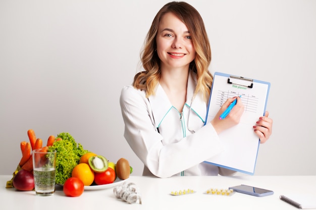 Joven médico dietista en la sala de consulta en la mesa con frutas y verduras frescas, trabajando en un plan de dieta