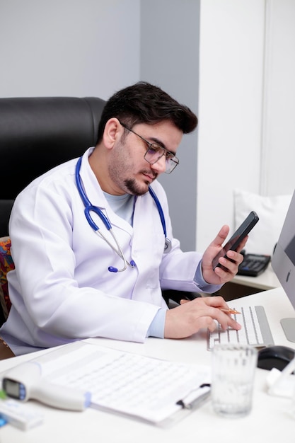 Un joven médico está sentado en la clínica sosteniendo el móvil en una mano y escribiendo algo en el teclado