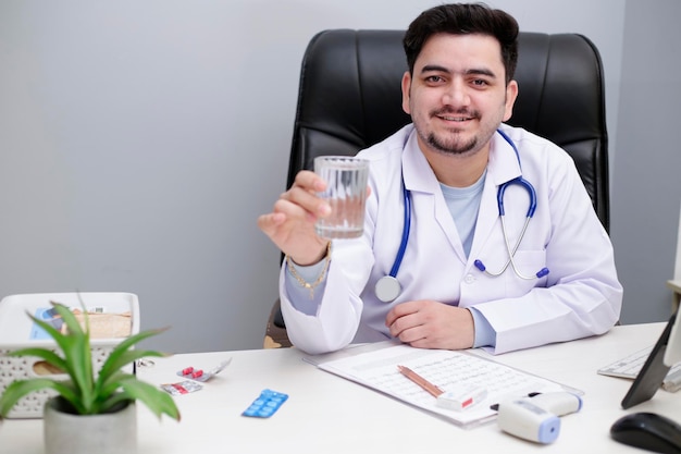 Un joven médico está sentado en la silla de la clínica y sostiene un vaso de agua en la mano