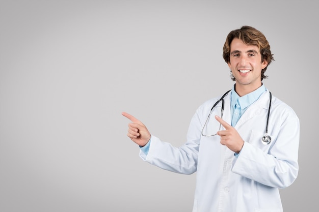 Un joven médico feliz apunta con los dedos al espacio vacío aislado en un fondo gris sonriendo a la cámara