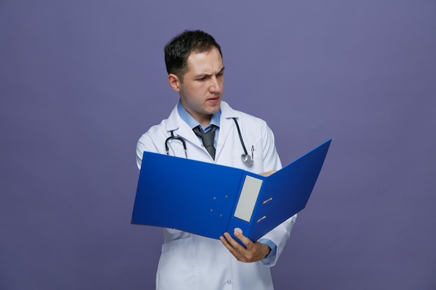 Joven médico frunciendo el ceño con bata médica y estetoscopio alrededor del cuello sosteniendo una carpeta apuntando con la mano leyendo de ella aislada en un fondo morado