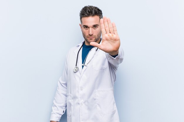 Joven médico guapo hombre de pie con la mano extendida que muestra la señal de stop