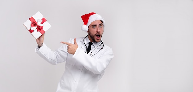 Joven médico guapo en uniforme blanco y sombrero de Santa Claus con presente