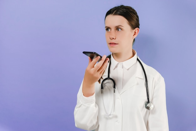 Joven médico habla con el teléfono inteligente para el comando de orden, posando aislado sobre fondo violeta de estudio con espacio de copia. Reconocimiento de voz biométrico, detección de voz en concepto de tecnología sanitaria