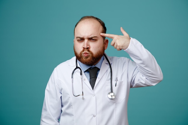 Un joven médico harto que usa un abrigo médico y un estetoscopio alrededor del cuello mirando a la cámara finge dispararse a sí mismo mostrando un gesto de suicidio aislado de fondo azul