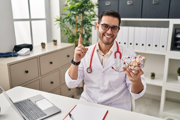 Joven médico hispano con barba sosteniendo dulces sorprendido con una idea o pregunta señalando con el dedo con la cara feliz número uno