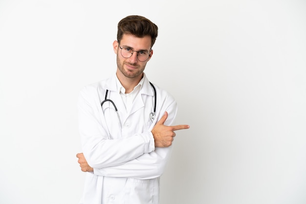 Joven médico hombre caucásico sobre aislado sobre fondo blanco vistiendo una bata de médico y apuntando hacia el lado