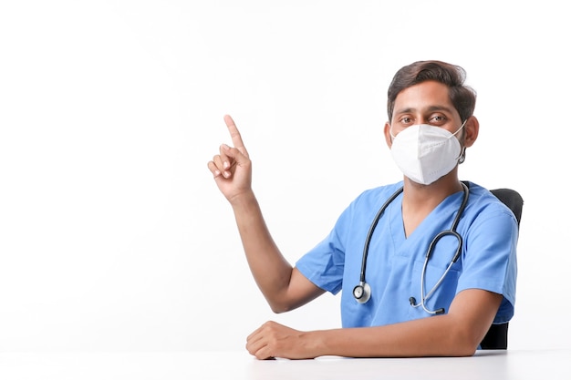 Joven médico indio con máscara y apuntando con la mano en la clínica.