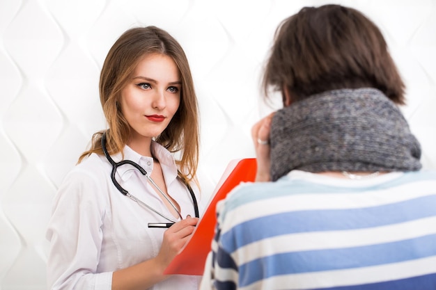 Joven médico inteligente con carpeta roja haciendo preguntas sobre enfermedad paciente chico guapo con bigote en cálida bufanda gris