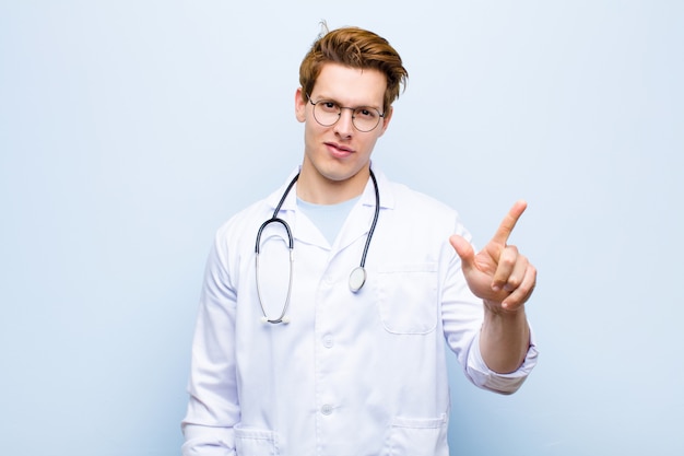 Joven médico jefe rojo sonriendo y mirando amigable, mostrando el número dos o segundo con la mano hacia adelante, contando hacia atrás