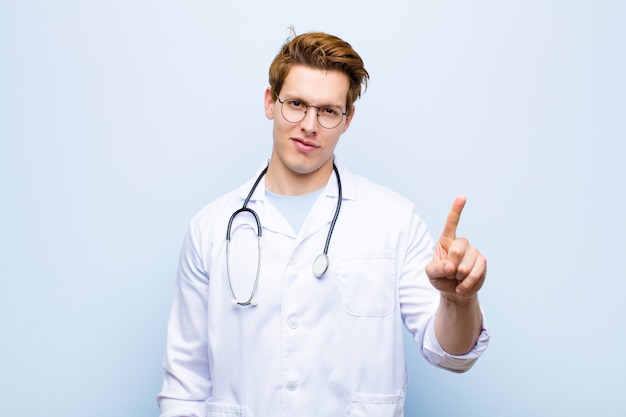 Joven médico jefe rojo sonriendo y mirando amigable, mostrando el número uno o primero con la mano hacia adelante, contando hacia atrás contra la pared azul