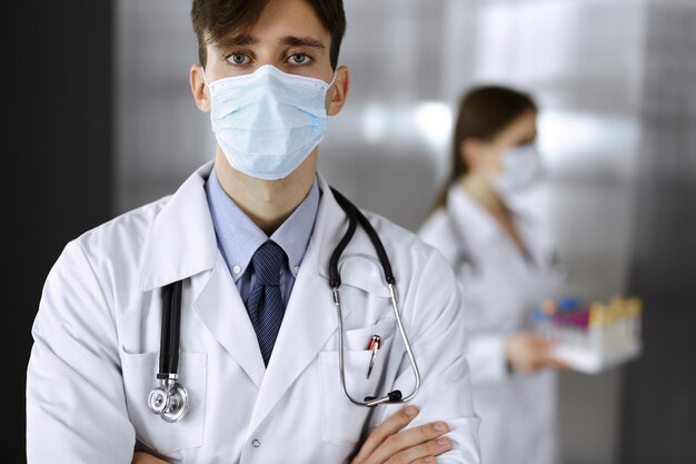 Joven médico masculino de pie con los brazos cruzados en la clínica. Médico inteligente con máscara protectora médica en un hospital moderno. Concepto de medicina y virus Corona.