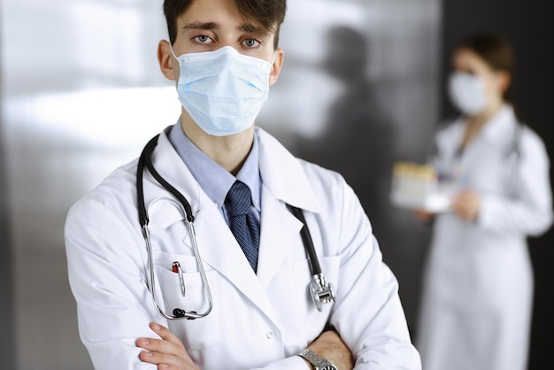 Joven médico masculino de pie con los brazos cruzados en la clínica. Médico inteligente con máscara protectora médica en un hospital moderno. Concepto de medicina y virus Corona.