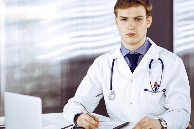 Joven médico masculino sentado y trabajando con el portapapeles del historial de medicamentos en la clínica en su lugar de trabajo. Médico rubio en el trabajo. Servicio médico perfecto. Concepto de medicina.