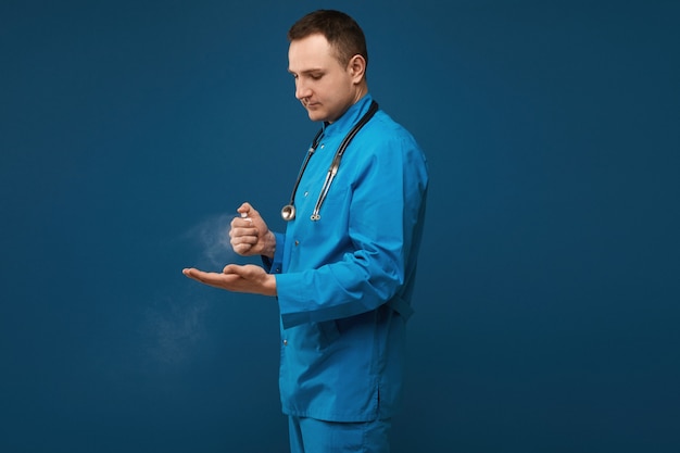 Joven médico masculino en uniforme médico azul con spray desinfectante para manos y posando en azul