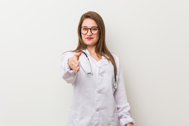 Joven médico mujer contra una pared blanca estirando la mano a la cámara en gesto de saludo