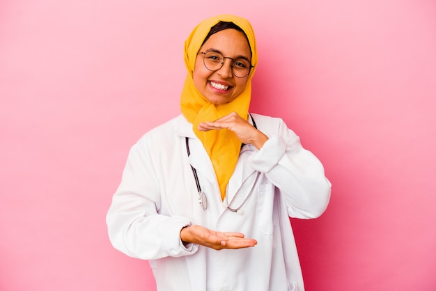 Joven médico mujer musulmana aislada en la pared rosa sosteniendo algo con ambas manos, presentación del producto.