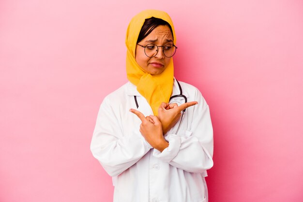 Joven médico mujer musulmana aislada sobre fondo rosa apunta hacia los lados, está tratando de elegir entre dos opciones.