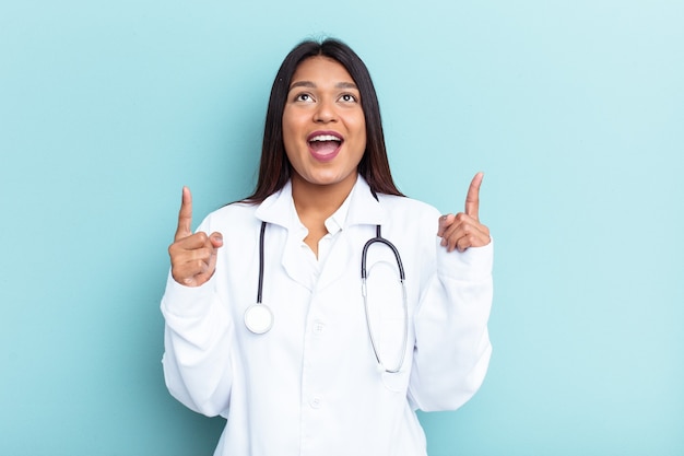 Joven médico mujer venezolana aislada sobre fondo azul apuntando al revés con la boca abierta.