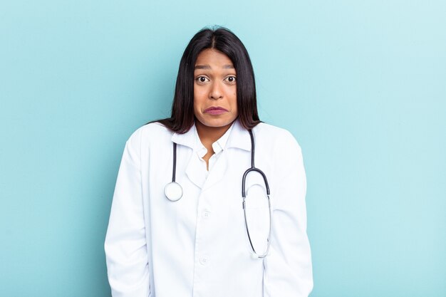 Joven médico mujer venezolana aislada sobre fondo azul se encoge de hombros y abre los ojos confundidos.