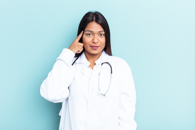 Joven médico mujer venezolana aislada sobre fondo azul señalando la sien con el dedo, pensando, centrado en una tarea.