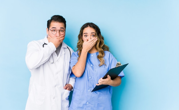 Foto joven médico pareja posando en un fondo azul sorprendido cubriendo la boca con las manos.