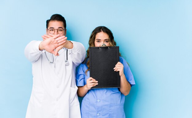 Joven médico pareja posando en una pared azul aislada haciendo un gesto de negación