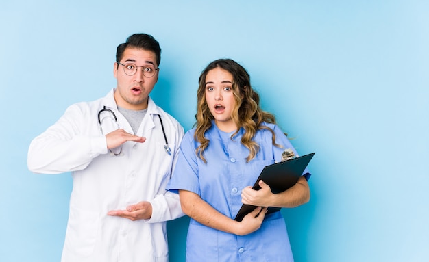 Joven médico pareja posando en una pared azul aislado sorprendido y sorprendido sosteniendo un espacio de copia entre las manos.