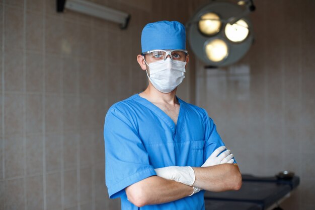 Joven médico profesional con uniforme azul, gafas y sombrero se encuentra en la sala de operaciones.